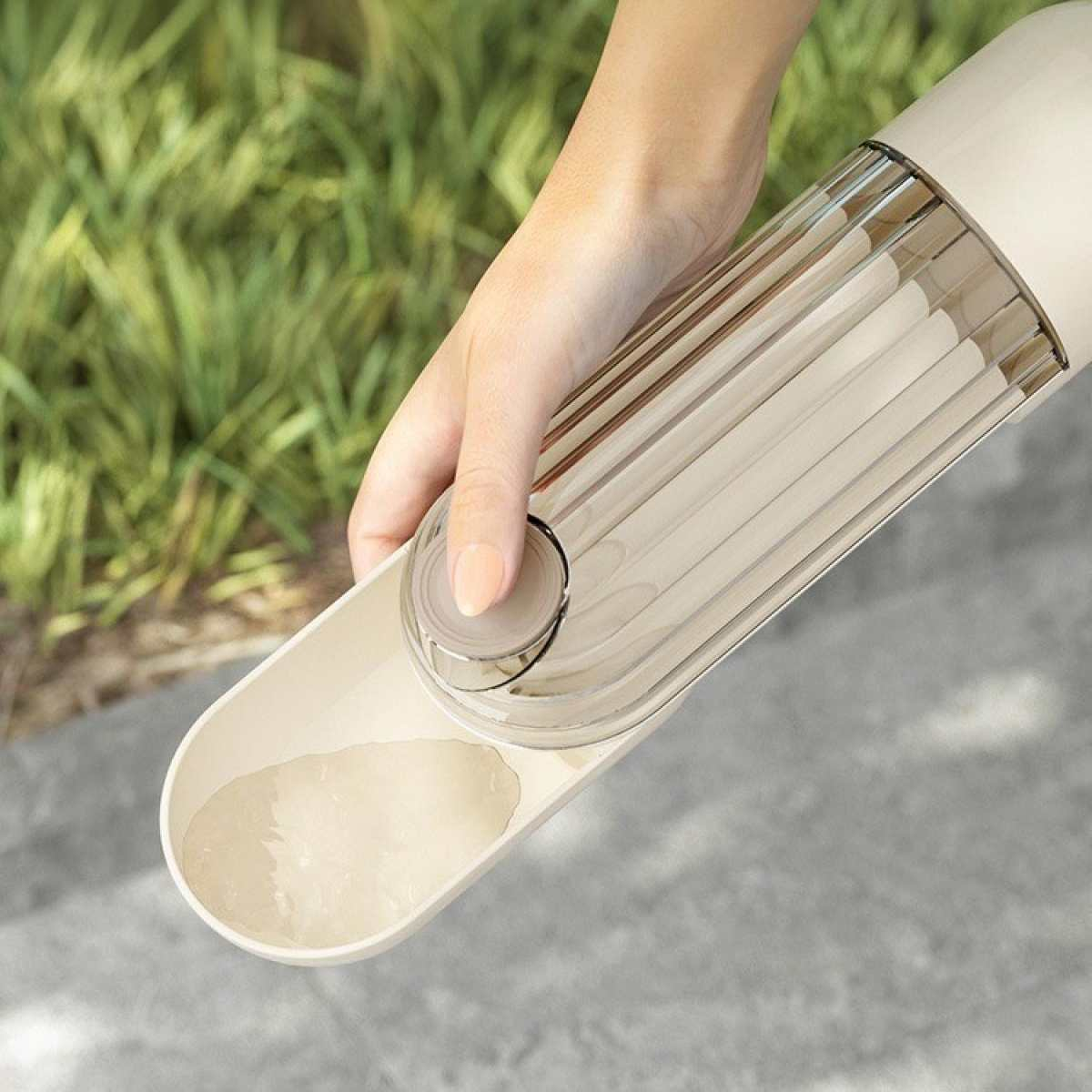 Eine Hand hält eine beige Hunde-Wasserflasche. Klares Wasser befindet sich im Trog. Grünes Gras ist im Hintergrund unscharf.