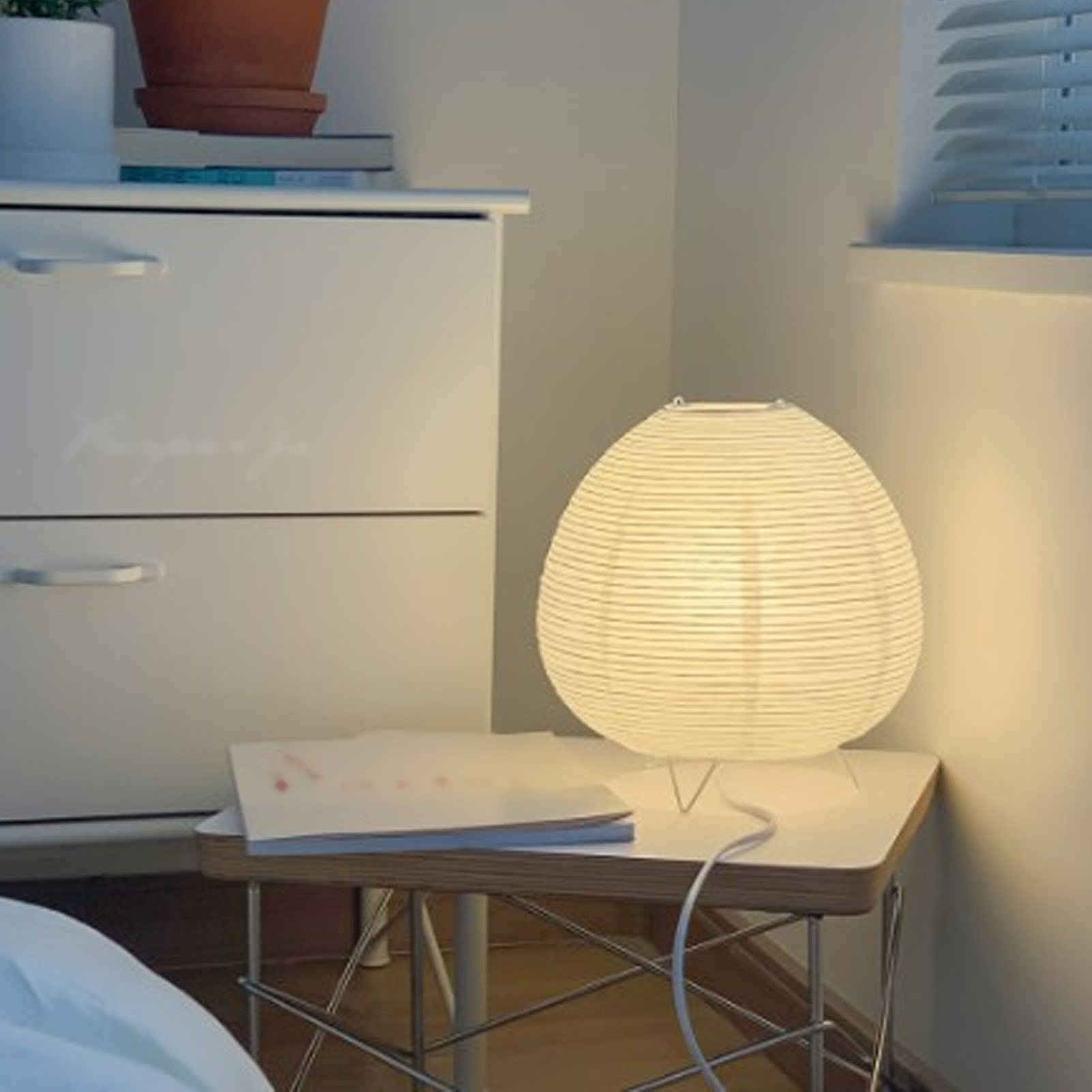 Eine Papierlampe steht auf einem Beistelltisch neben einer weißen Kommode. Ein Fenster mit Jalousien ist sichtbar.
