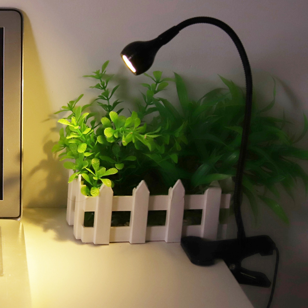 Eine schwarze Schreibtischlampe beleuchtet eine kleine Pflanze in einem weißen Zäunchen. Die Lampe ist am Schreibtisch befestigt.