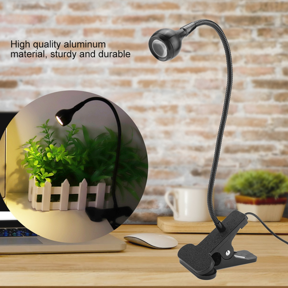 Eine schwarze Schreibtischlampe mit flexiblem Hals. Sie ist an einem Schreibtisch befestigt. Ein kleiner Blumentopf ist sichtbar.