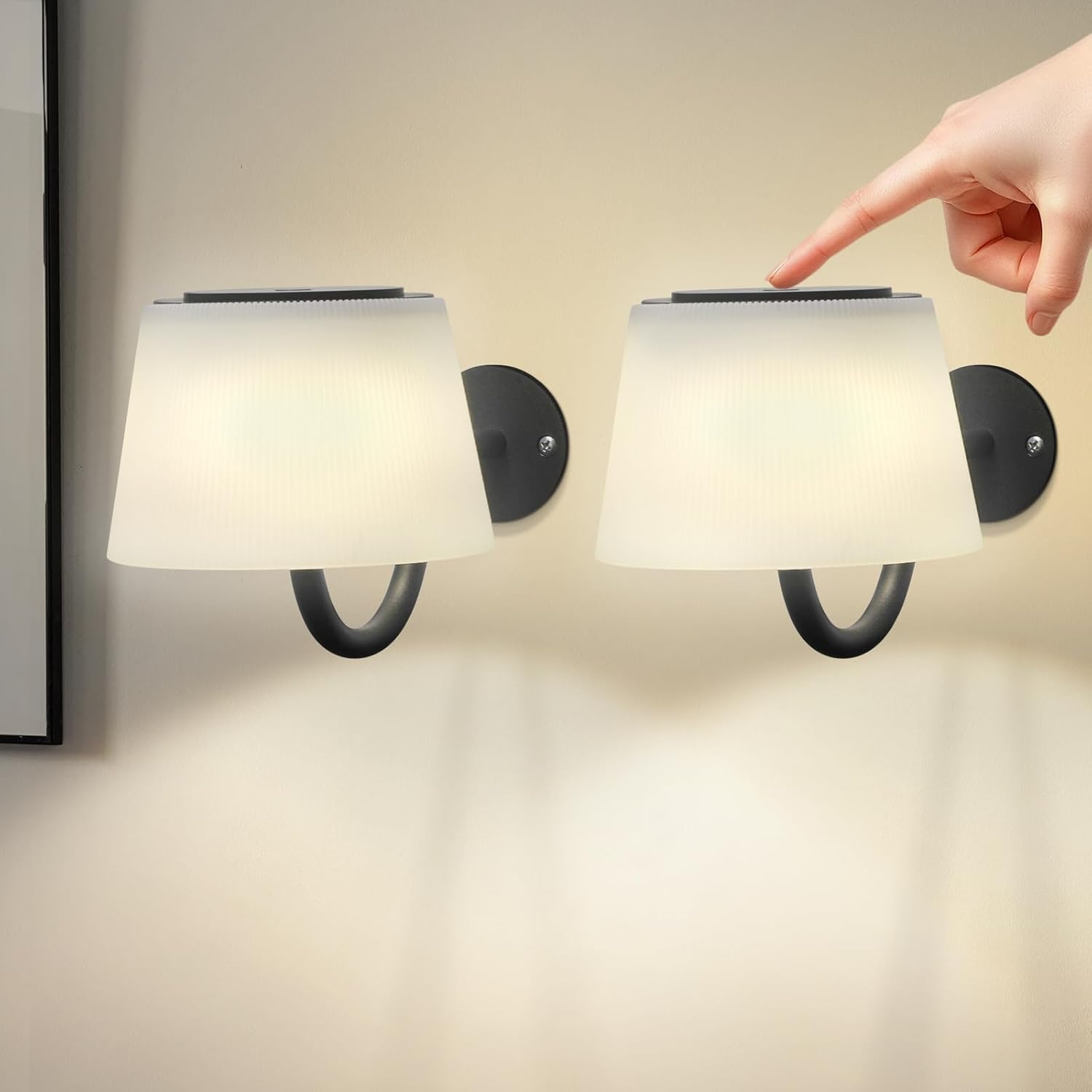 Due lampade da parete con paralumi bianchi su un muro beige. Una mano tocca la parte superiore di una lampada. Cornici nere a sinistra.