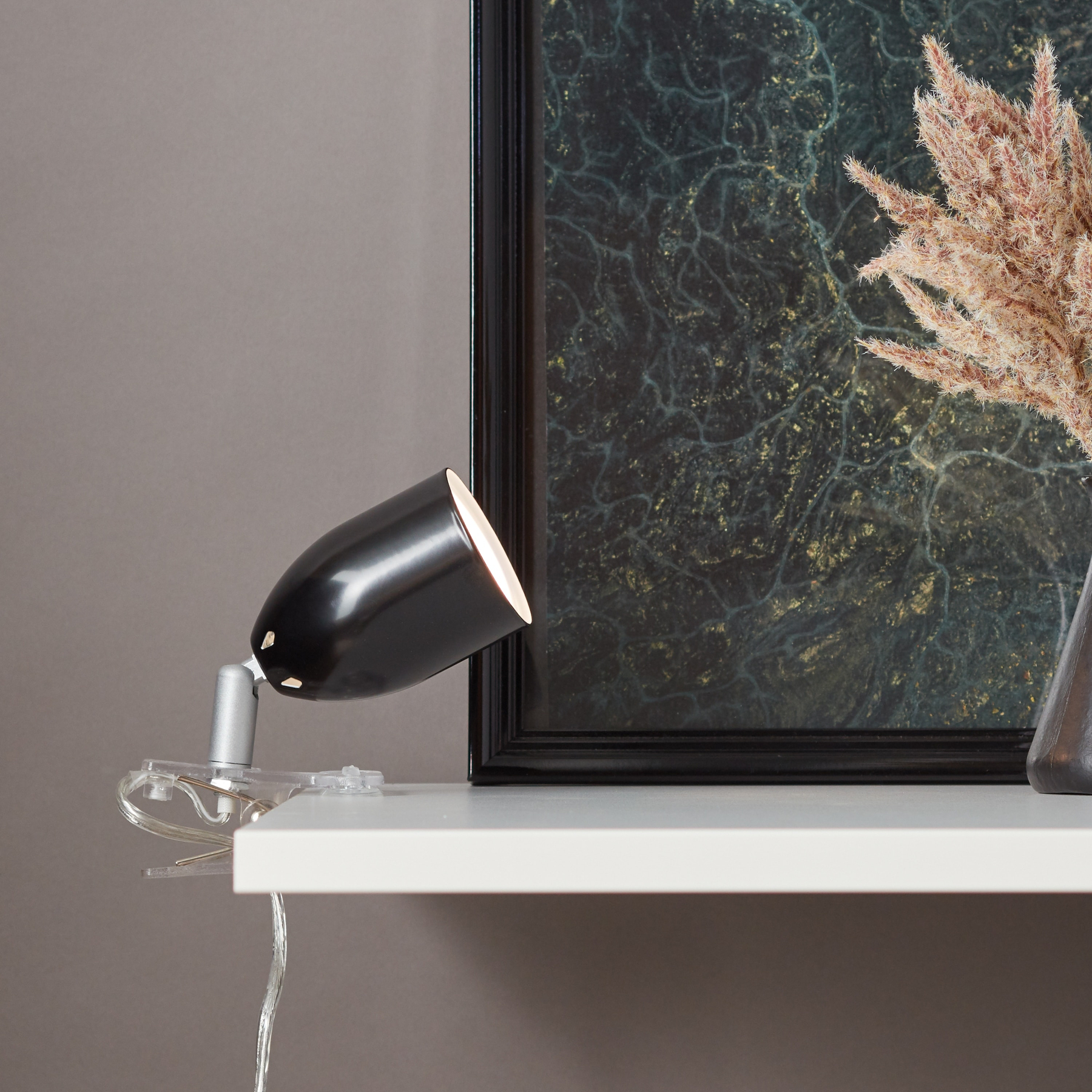 Schwarze Lampe auf weißem Regal. Dahinter ein gerahmtes abstraktes Kunstwerk und eine Vase mit Trockenblumen.