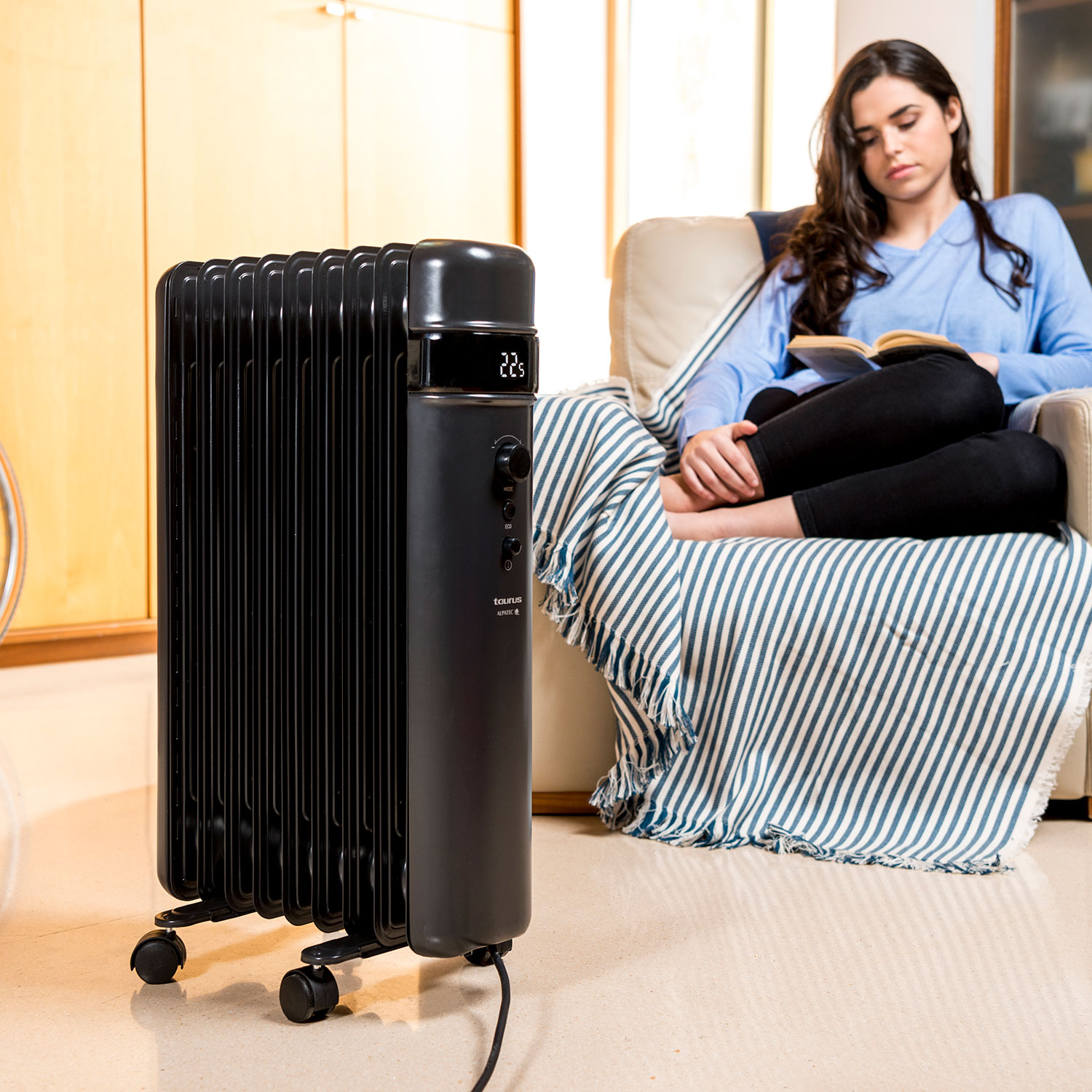 Ein schwarzer Ölradiator steht in der Nähe einer Frau, die auf einer Couch liest. Die Frau liegt unter einer gestreiften Decke.