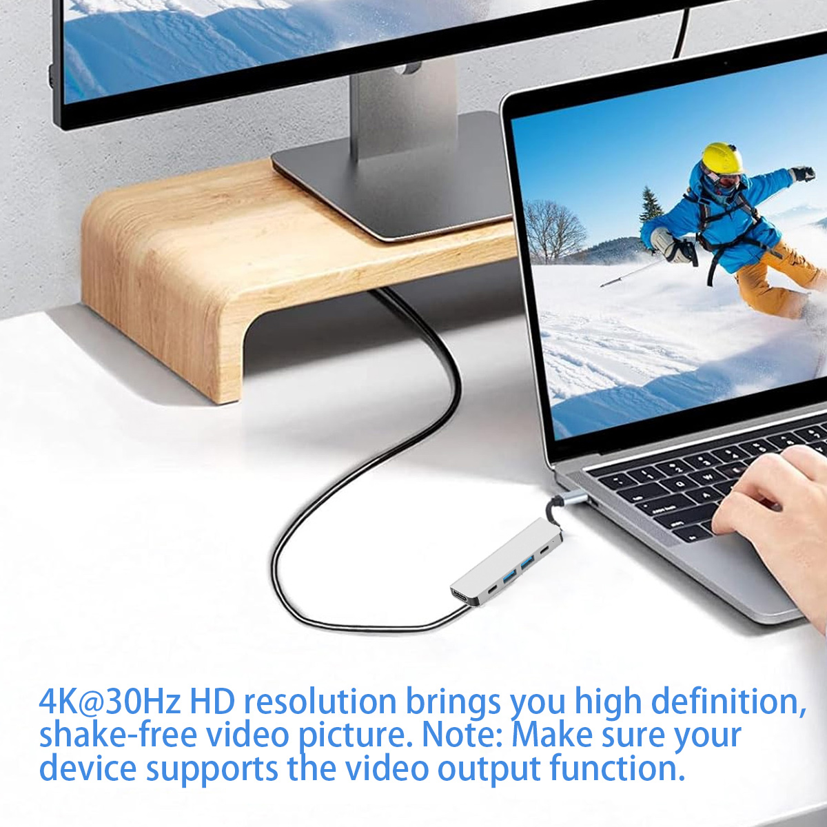 Ein Laptop zeigt einen Skifahrer. Ein Hub ist mit dem Laptop verbunden. Ein Monitor steht auf einem Holzständer. Eine Hand berührt den Laptop.