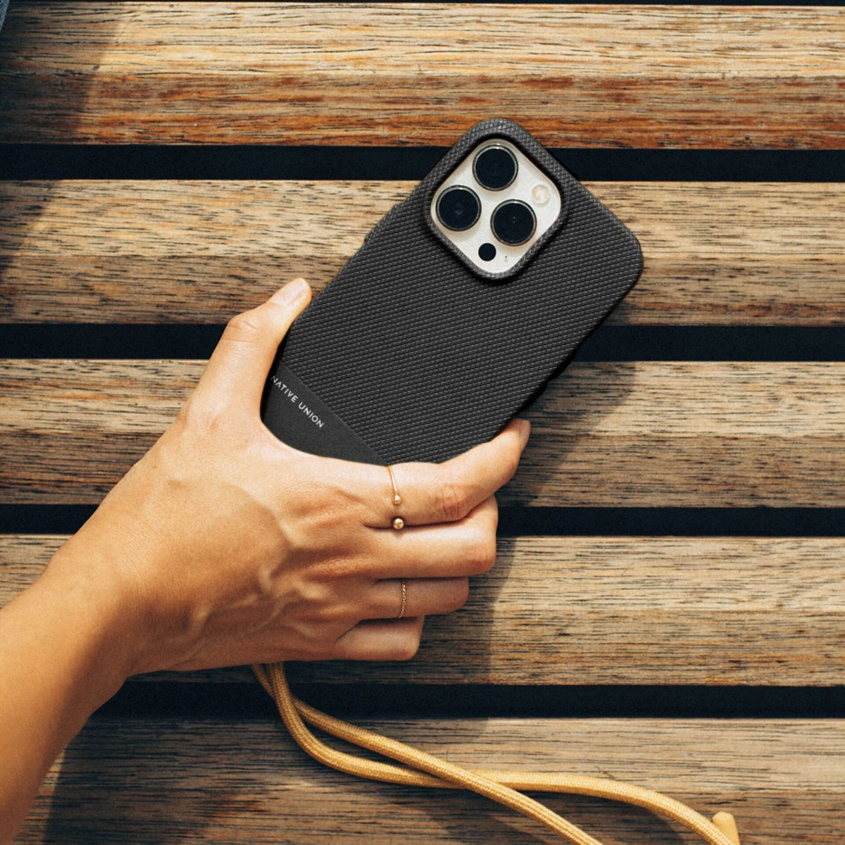 Una mano tiene un telefono nero con una custodia, appoggiato su assi di legno. Una corda dorata è attaccata al telefono. Anelli adornano le dita.