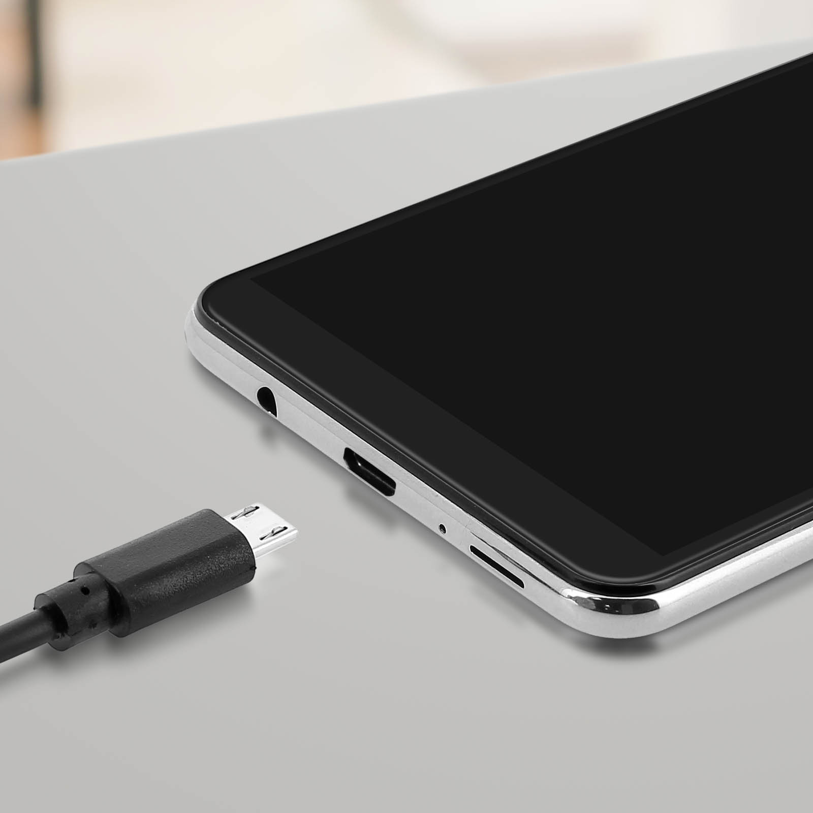 Uno smartphone nero è appoggiato su una superficie grigio chiaro accanto a un cavo di ricarica nero. Lo schermo è nero.