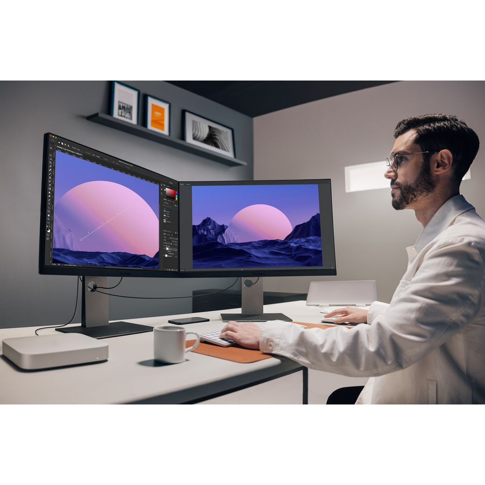 Hombre trabajando en monitores duales, mostrando un paisaje surrealista. Usa gafas y bata blanca, en una oficina moderna.