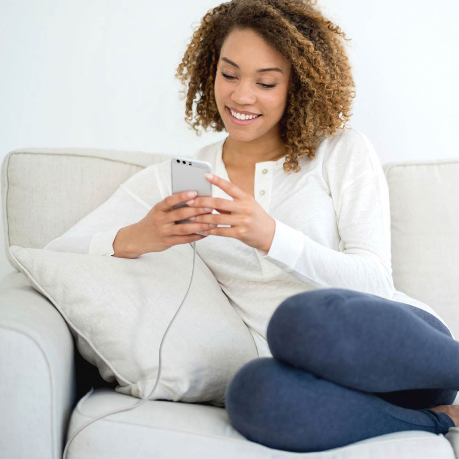 Una donna con i capelli ricci è seduta su un divano, sorridendo. Tiene in mano un telefono, indossa una camicia bianca e pantaloni blu.