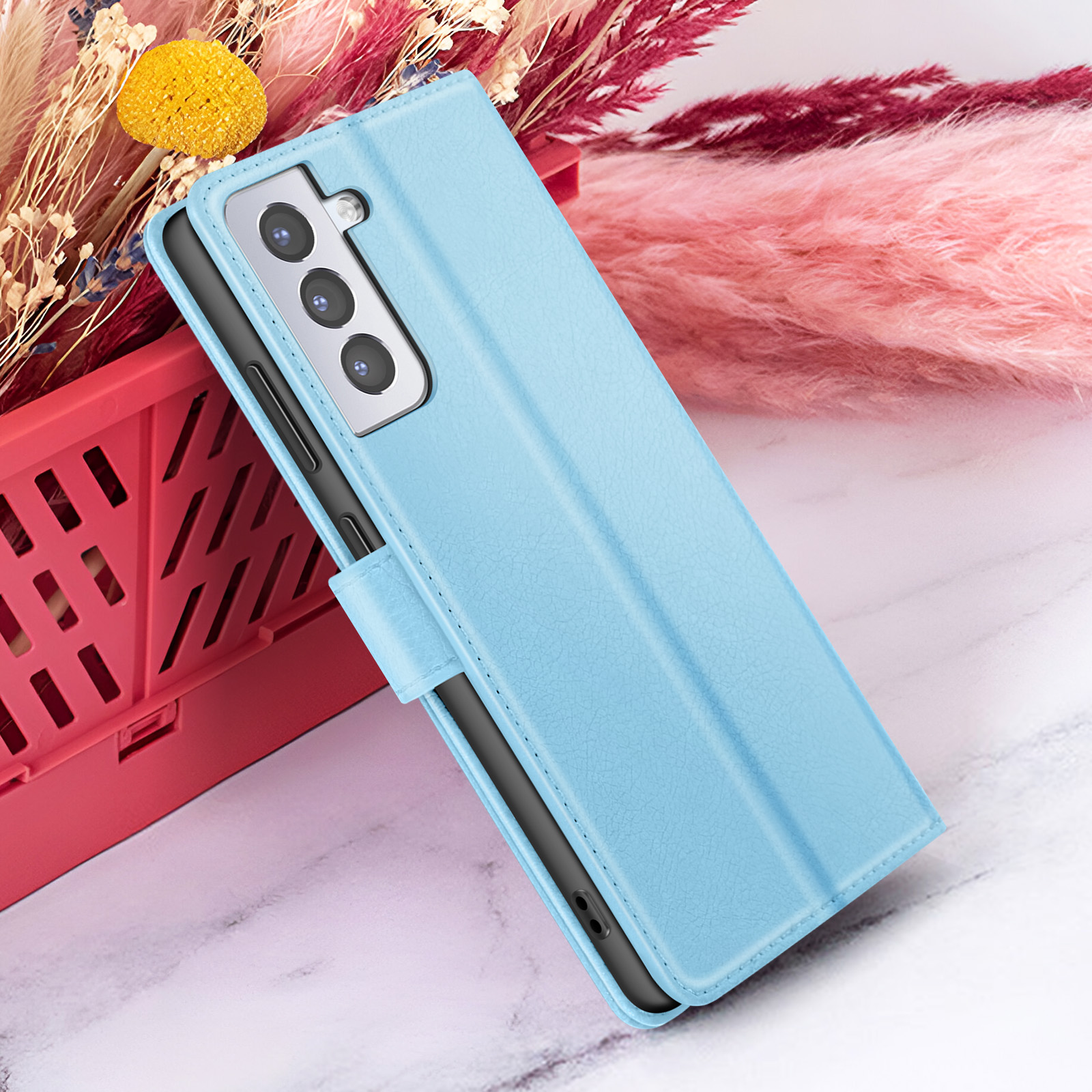 Un étui de téléphone bleu clair est présenté, appuyé contre un conteneur rouge avec des fleurs séchées en arrière-plan.