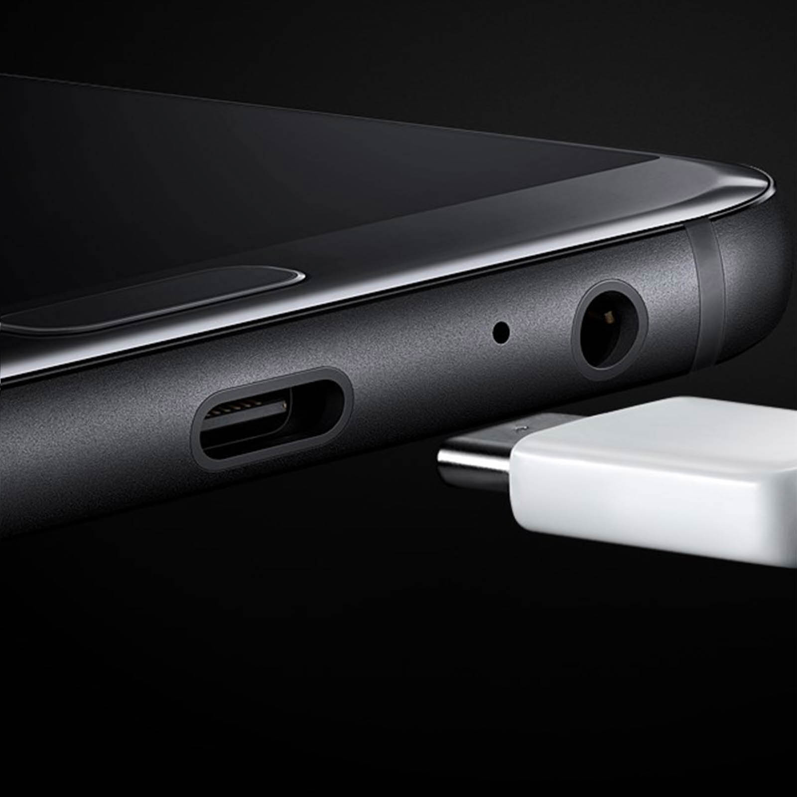 Primo piano di un telefono nero con adattatore bianco collegato. Mostra le porte: USB-C, jack per cuffie. Sfondo nero.