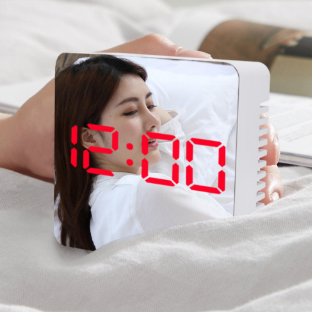 Eine Frau spiegelt sich in einer Digitaluhr. Die Uhr zeigt 12:00 in Rot. Die Frau scheint im Bett auf weißen Laken zu liegen.