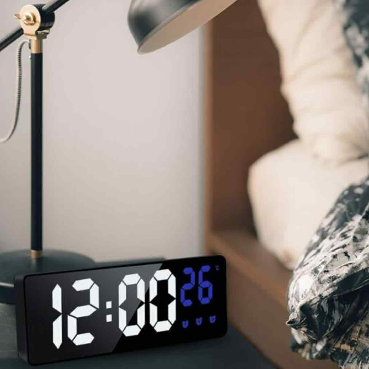 Eine Digitaluhr zeigt 12:00:26 an. Im Hintergrund befindet sich eine schwarze Schreibtischlampe. Rechts ist ein Bett mit Kissen teilweise zu sehen.