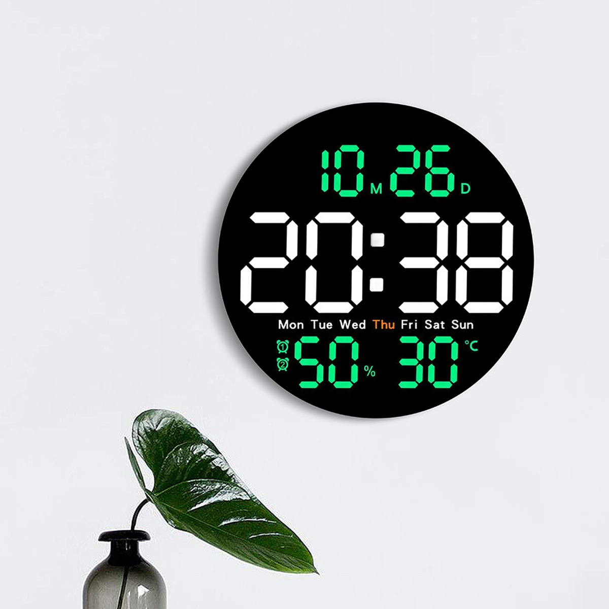 Eine Digitaluhr zeigt Uhrzeit und Datum an. Sie zeigt 20:38 und 10.26. Die Luftfeuchtigkeit beträgt 50 % und die Temperatur 30 °C. Eine Pflanze ist sichtbar.