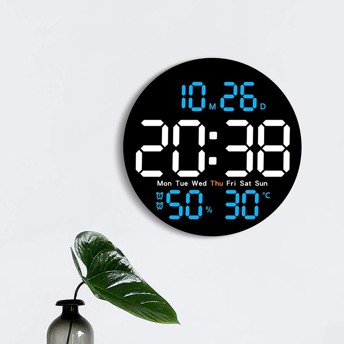 Eine schwarze Digitaluhr zeigt Zeit, Datum und Luftfeuchtigkeit an. Eine grüne Pflanze in einer Vase befindet sich im Vordergrund.