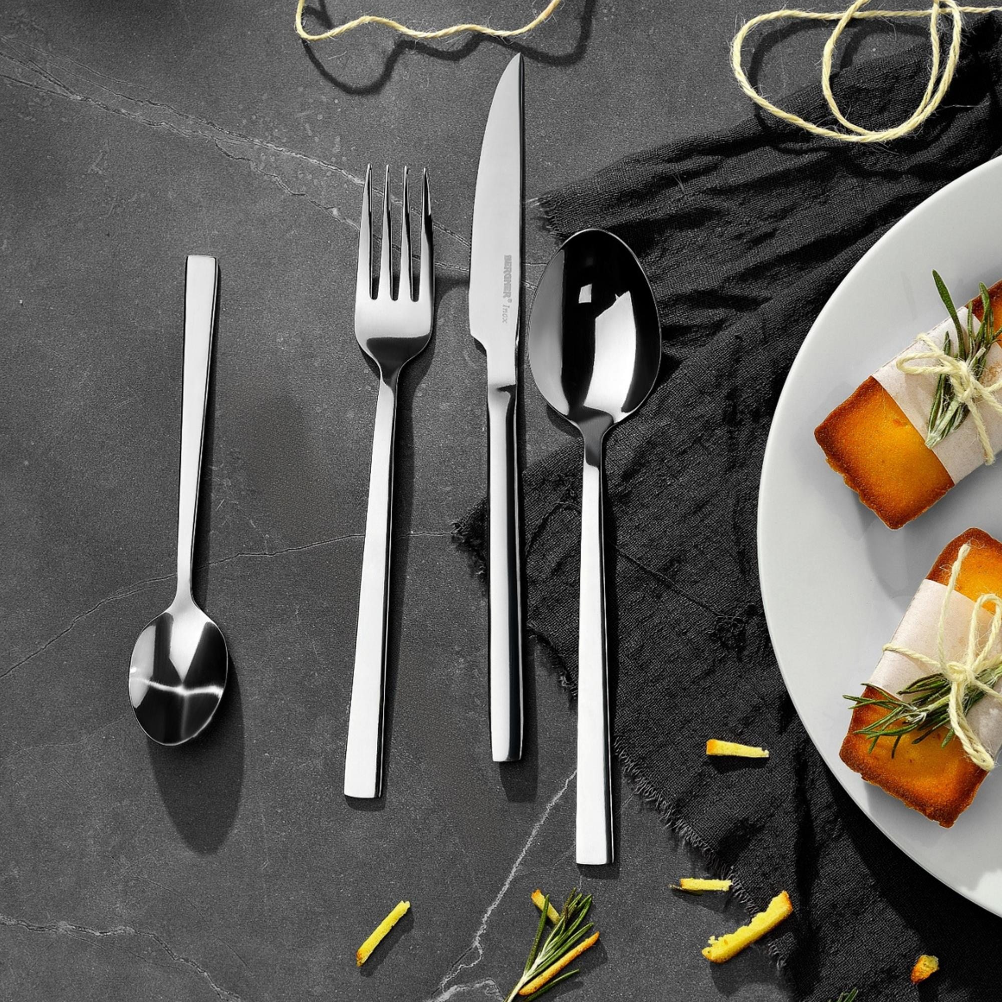 Juego de cubiertos plateados junto a un plato con pasteles envueltos, sobre una superficie oscura. Incluye tenedor, cuchara, cuchillo y postres.