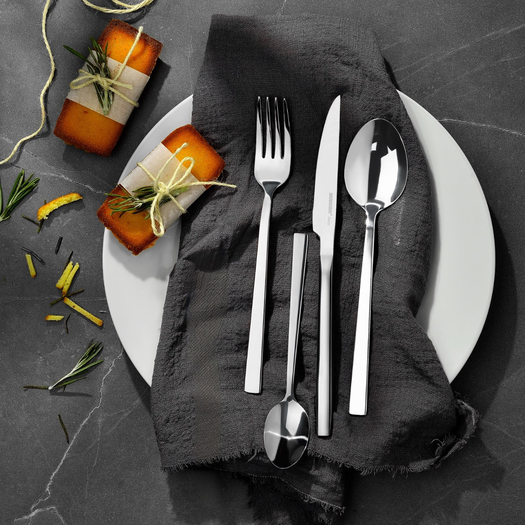 Decoración de mesa con tenedor, cuchillo, cuchara en un plato, servilleta oscura y dos pasteles envueltos con romero.