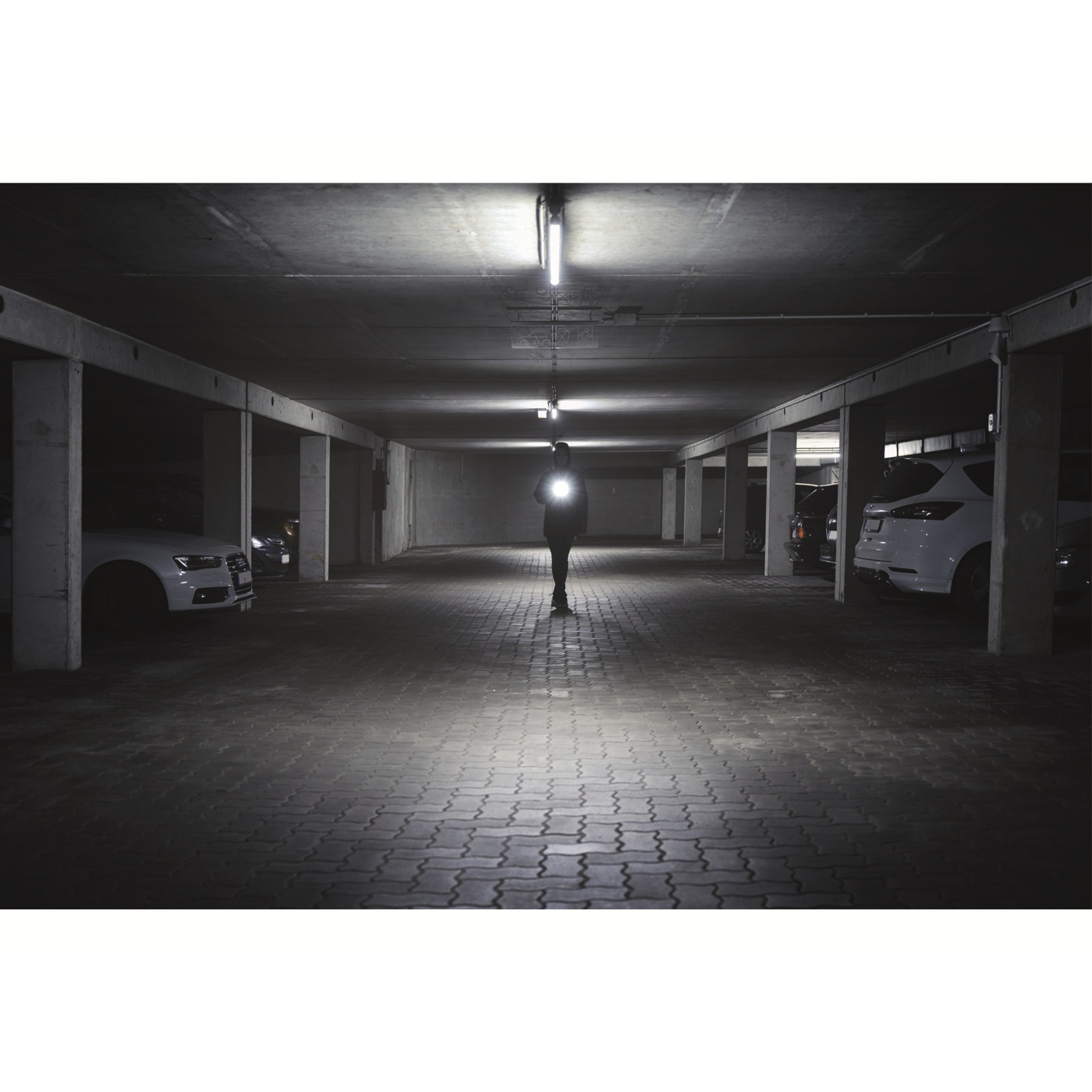 Eine Person hält eine Taschenlampe in einer dunklen Parkgarage. Autos stehen auf beiden Seiten. Der Boden hat ein Ziegelmuster.