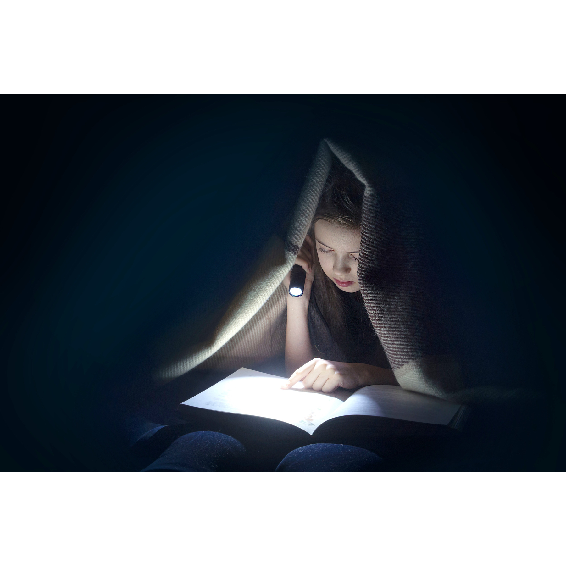 Dziewczyna czyta książkę pod kocem, oświetlona latarką. Scena jest słabo oświetlona.