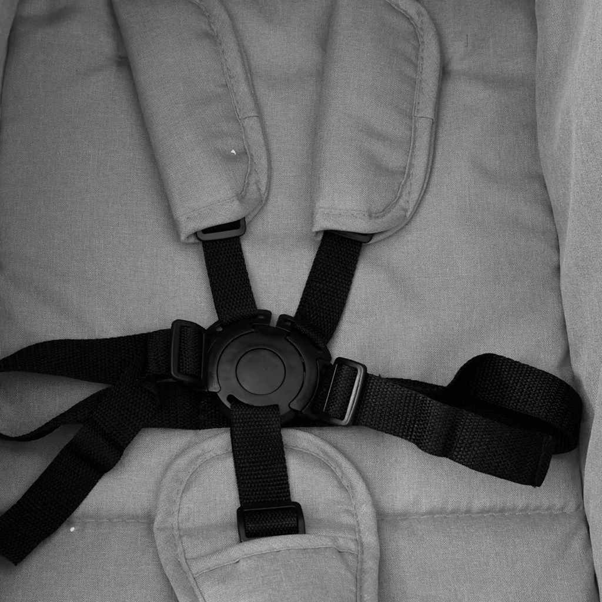 Nahaufnahme eines Kinderwagen-Gurts. Schwarze Gurte und Schnalle vor hellgrauem Stoffhintergrund. Der Gurt scheint unbenutzt.