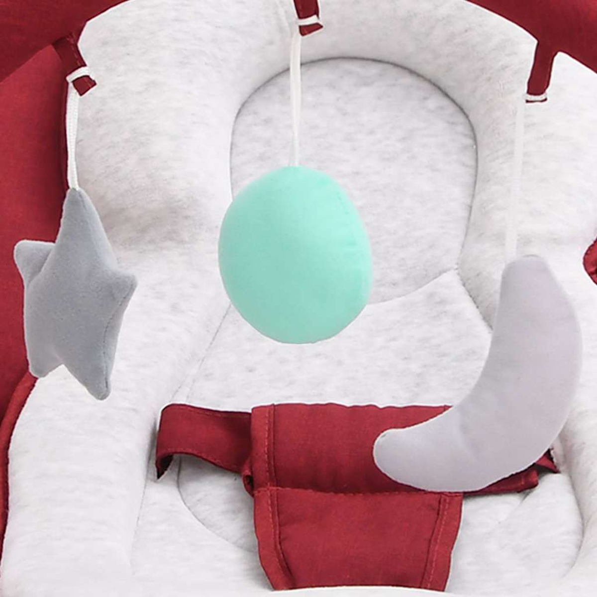 Eine Babywippe mit hängendem Spielzeug: ein Stern, ein Mond und ein grüner Ball. Die Wippe ist rot und grau.