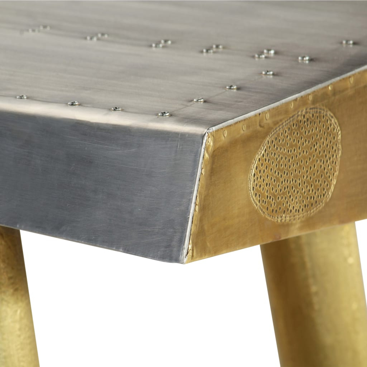 Nahaufnahme eines Metalltisches. Die Oberseite ist silberfarben mit Nieten. Die Seite ist goldfarben mit einem ovalen Lochmuster. Goldene Beine stützen den Tisch.
