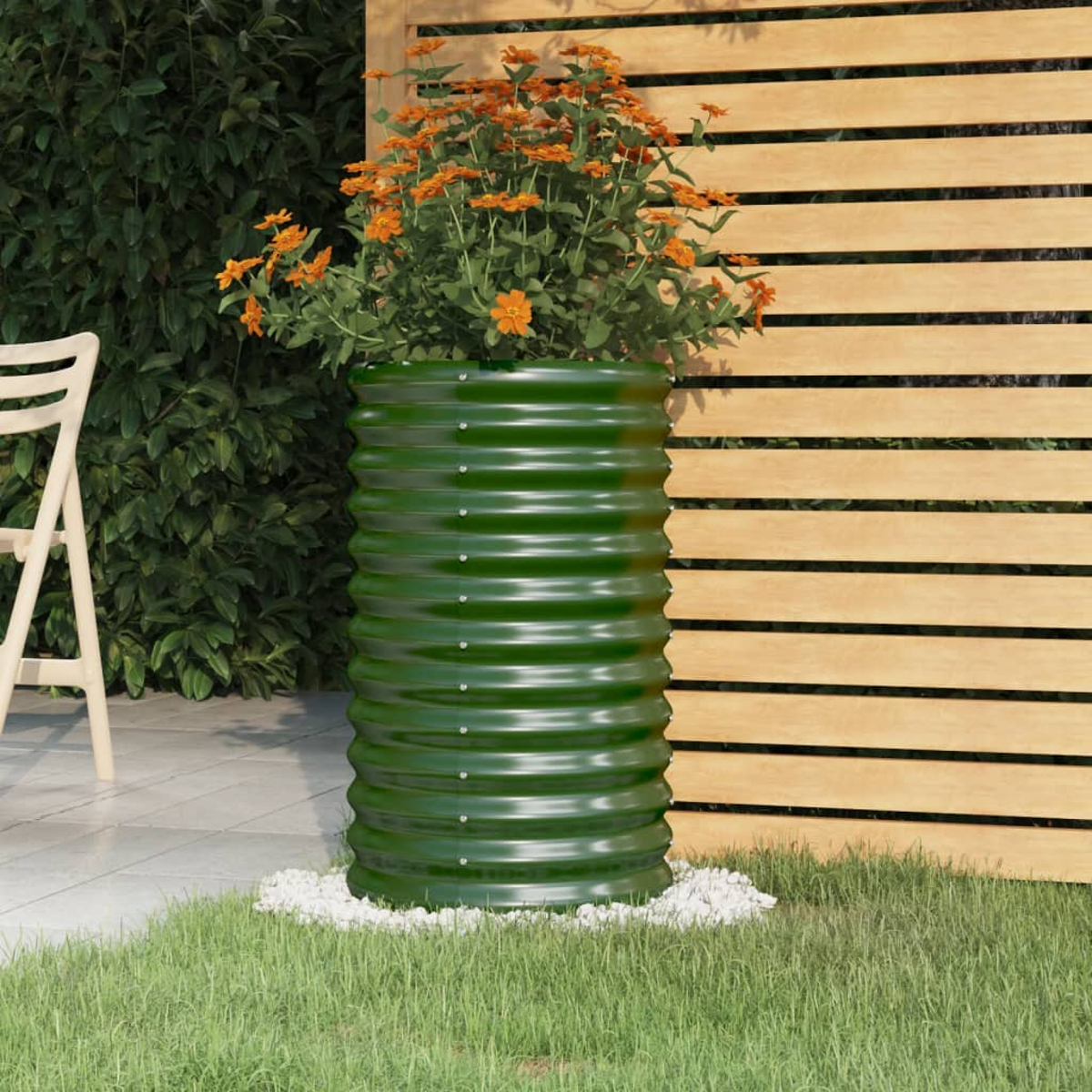 Ein grüner Wellenpflanzer mit orangefarbenen Blumen steht auf Steinen vor einem Holzzaun und Gras.