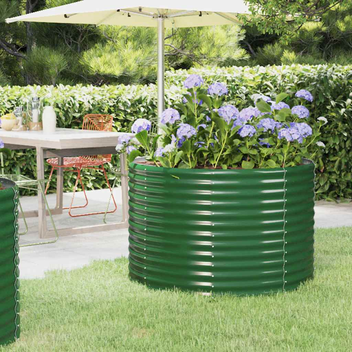Grünes Metall-Gartenbeet mit lila Blumen. Tisch mit Sonnenschirm im Hintergrund. Grünes Gras und Hecke umgeben die Szene.