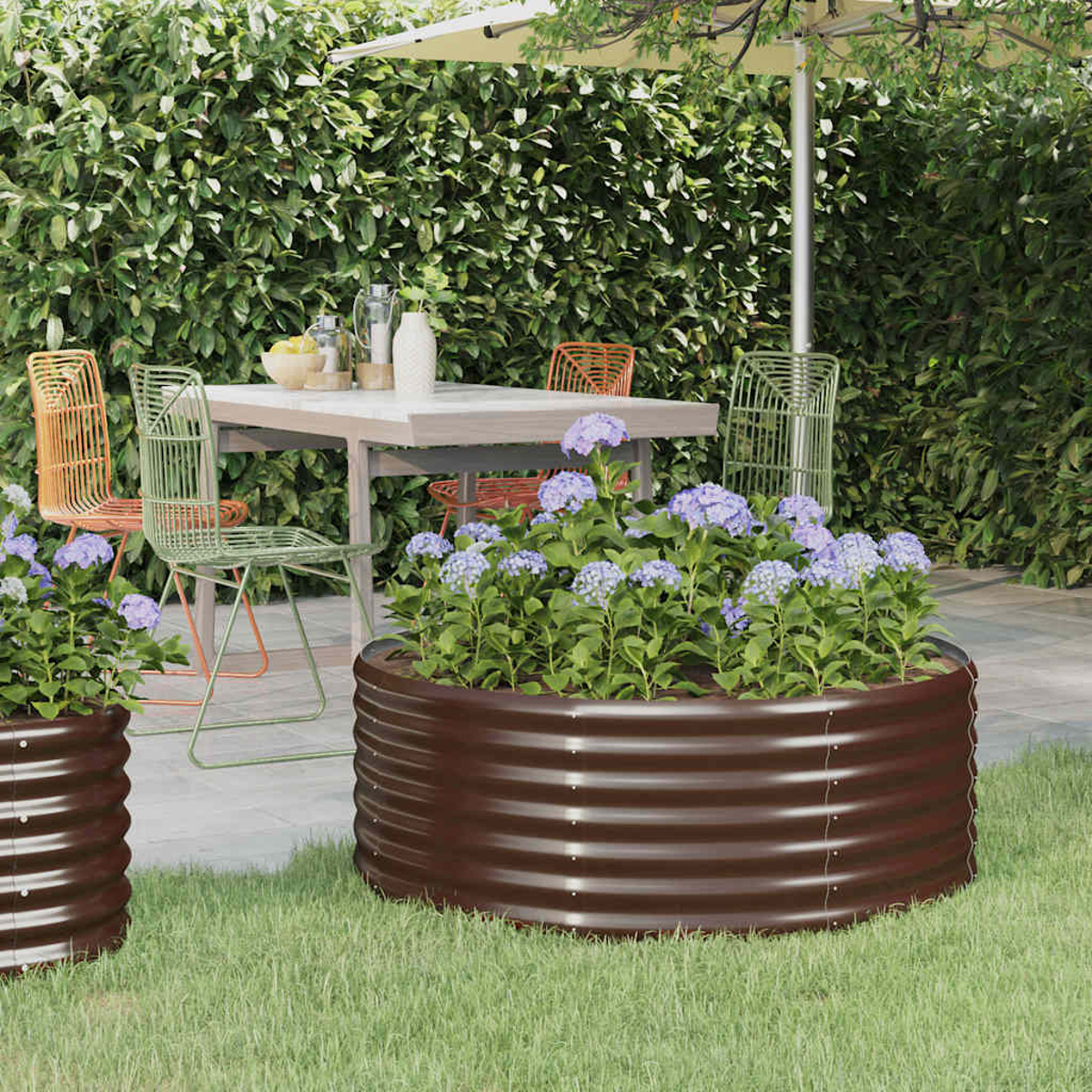 Gartenszene mit braunen Metallpflanzgefäßen mit lila Blumen, einem Tisch mit Stühlen und einem grünen Heckenhintergrund.
