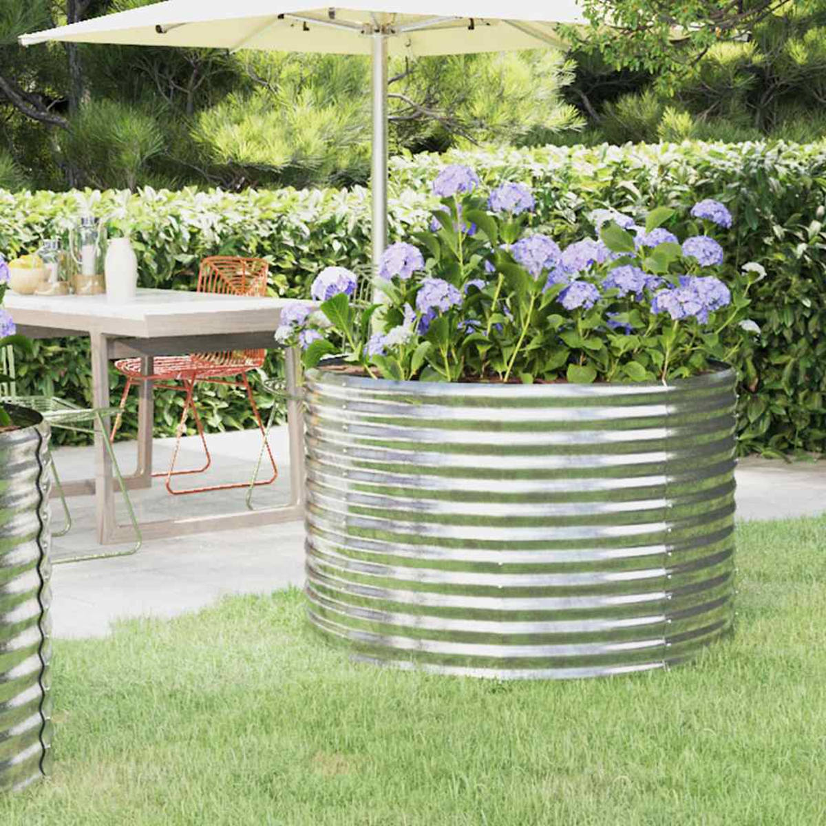 Eine Gartenszene mit einem Metallpflanzgefäß mit lila Blumen, einem Tisch, Stühlen und einem Sonnenschirm auf grünem Gras.