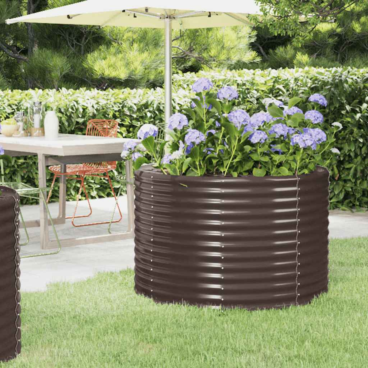 Ein brauner Metallpflanzer mit blauen Blumen steht auf Gras. Im Hintergrund sind ein Sonnenschirm und ein Tisch.