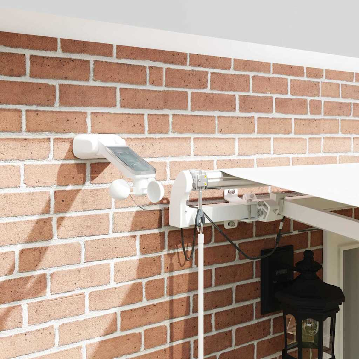 Eine Backsteinmauer mit einer weißen Wetterstation, Markise und einer schwarzen Lampe. Die Wetterstation hat ein Solarpanel und zwei weiße Kugeln.