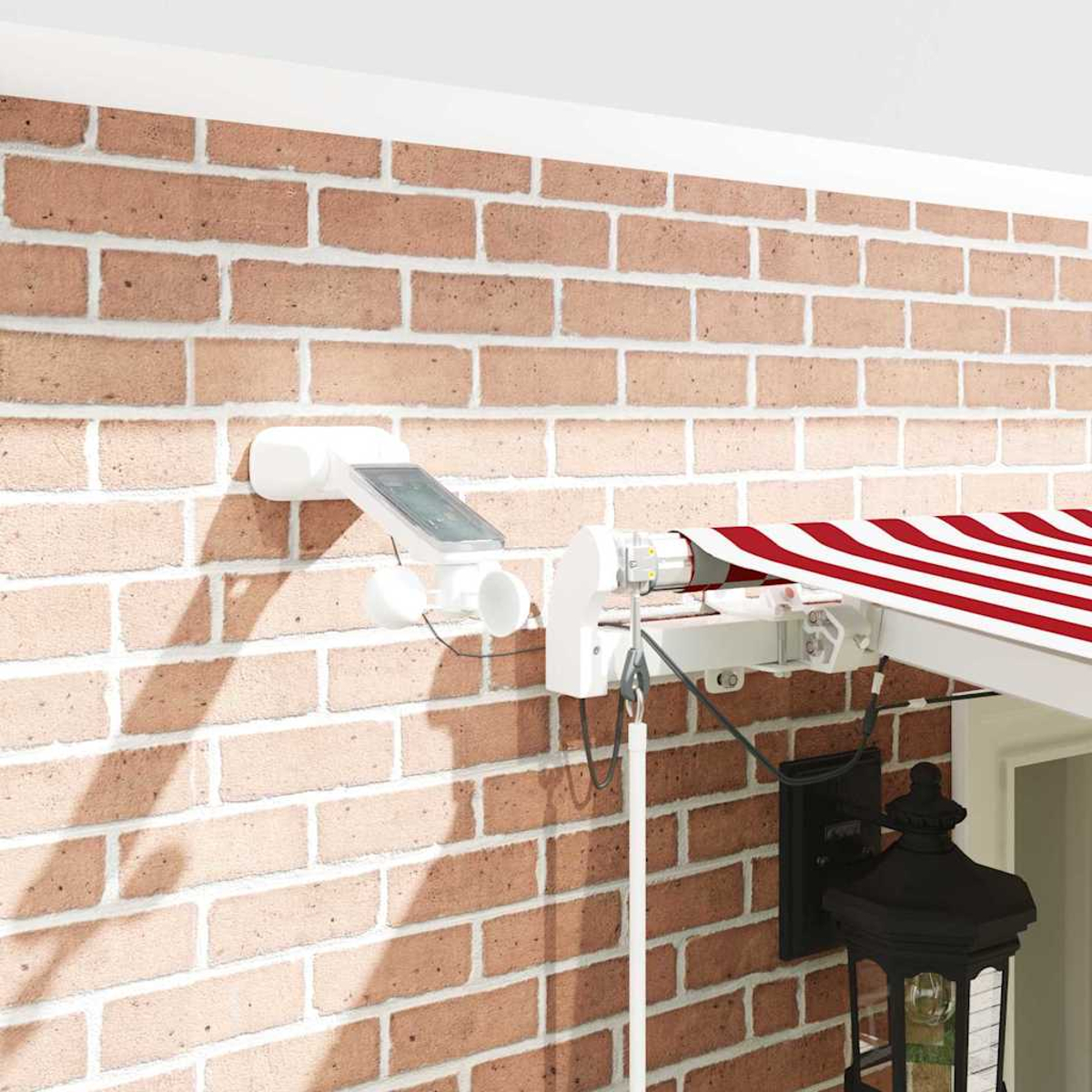 Ziegelmauer mit Markise und Sensor. Weiß-rot gestreifte Markise. Weißer Sensor mit Solarpanel. Schwarze Laterne.