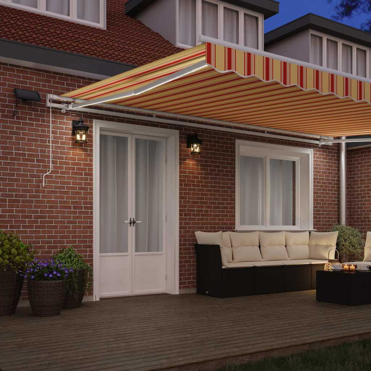 Ein Backsteinhaus mit gestreifter Markise. Eine Terrasse hat ein Sofa und einen Tisch. Blumentöpfe vor einer weißen Tür. Lichter sind an.