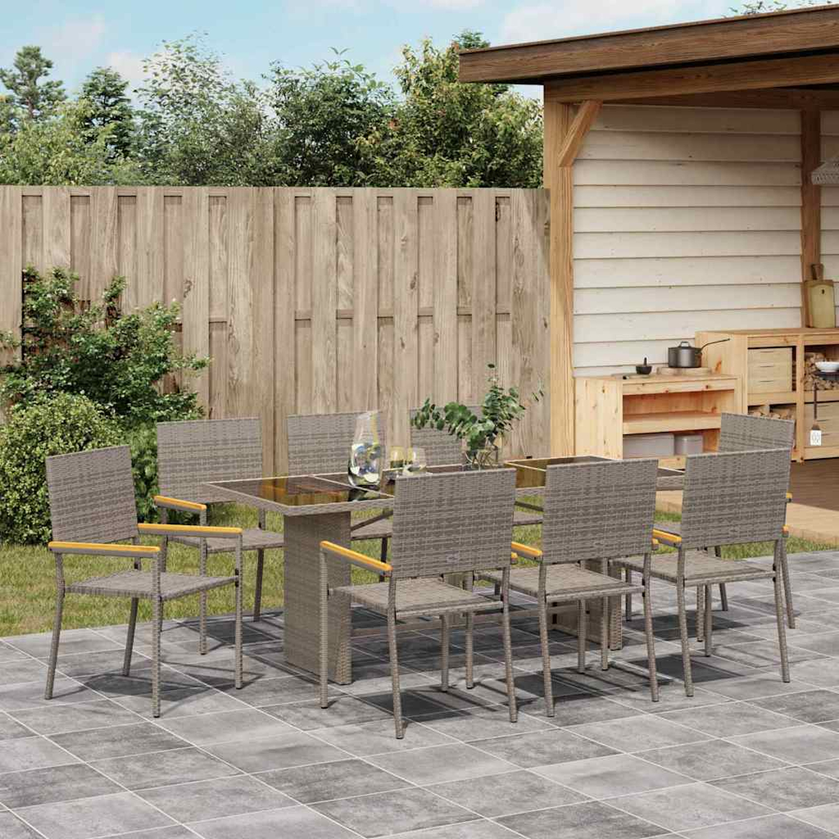 Essgruppe im Freien. Korbstühle um einen Tisch mit Glasplatte auf einer Terrasse. Holzzaun und Struktur mit Grün im Hintergrund.
