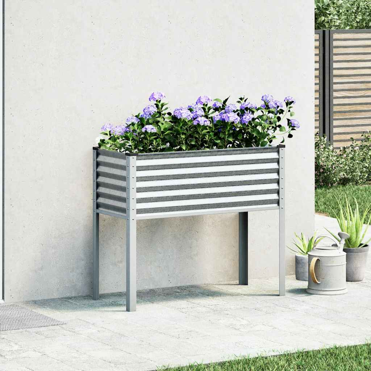 Una fioriera rialzata in metallo con fiori viola contro un muro, con un annaffiatoio e piante vicine.