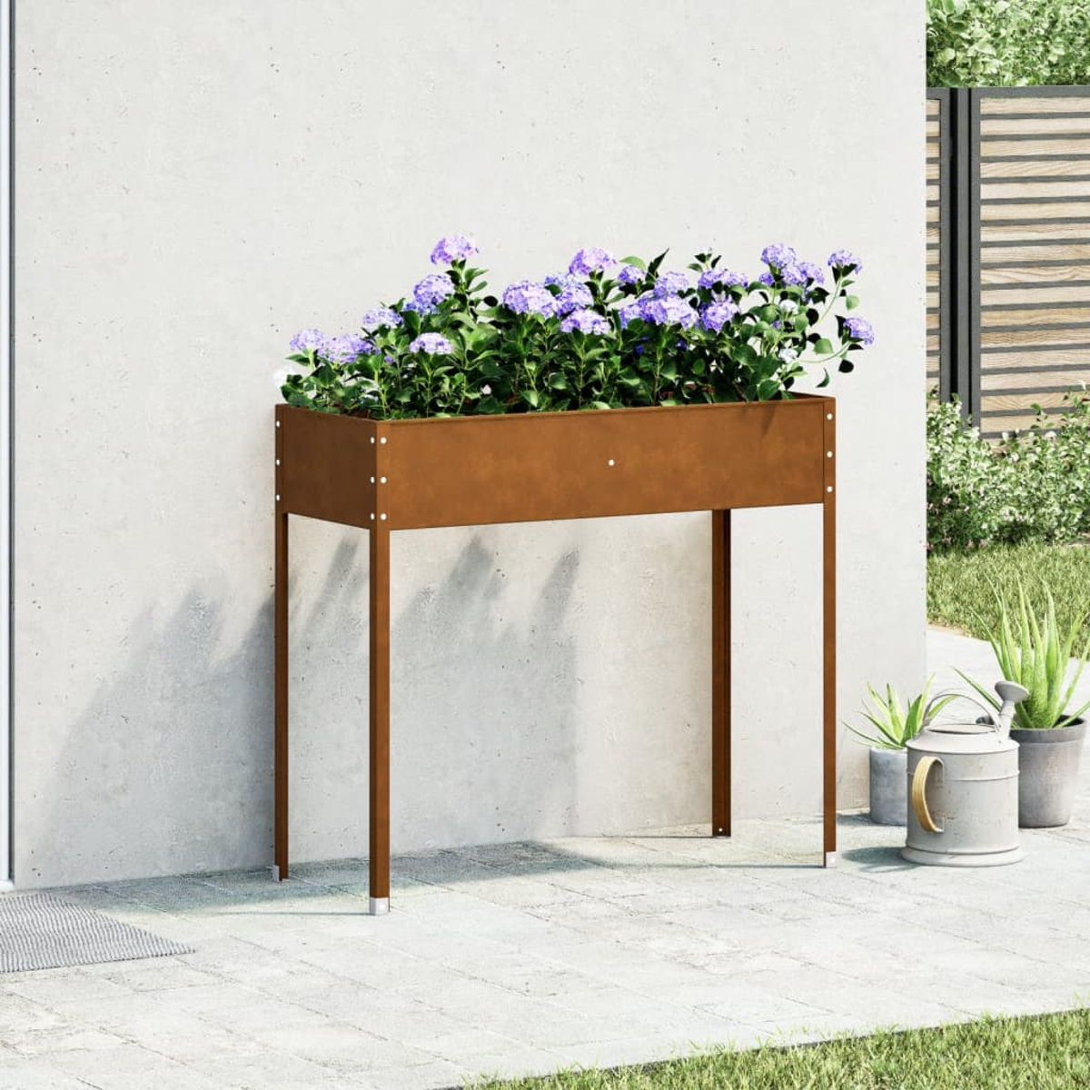 Una fioriera rialzata con fiori viola si trova su un patio. Un annaffiatoio e piante in vaso sono vicini, contro un muro.