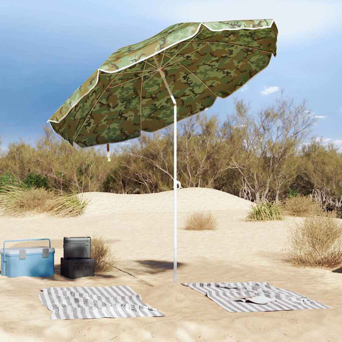 Scena sulla spiaggia con ombrellone mimetico, frigorifero e coperte sulla sabbia, con cespugli e cielo blu sullo sfondo.