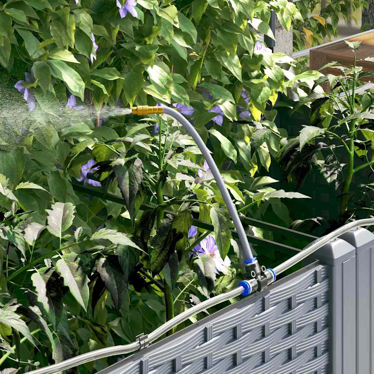 Un irrigatore spruzza acqua su piante verdi con fiori viola. L'irrigatore è attaccato a una recinzione grigia.