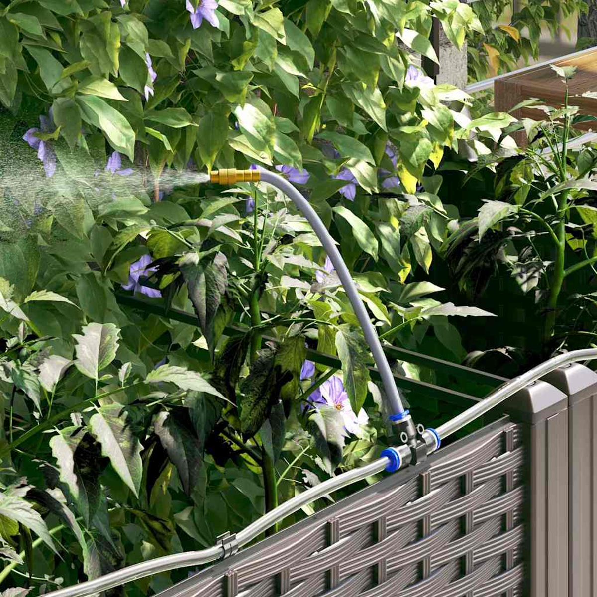Un tubo da giardino spruzza acqua su piante verdi e fiori viola. Il tubo è collegato a una recinzione marrone.
