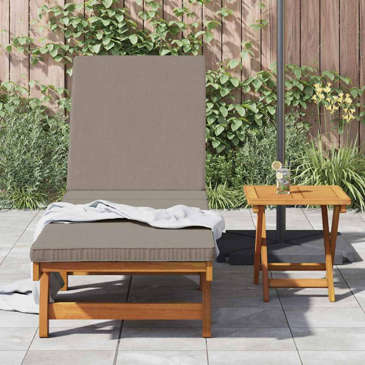 Eine Szene auf der Terrasse mit Liegestuhl, Beistelltisch mit Getränk und Holzzaun mit Pflanzen. Stuhl hat ein Handtuch und Kissen.