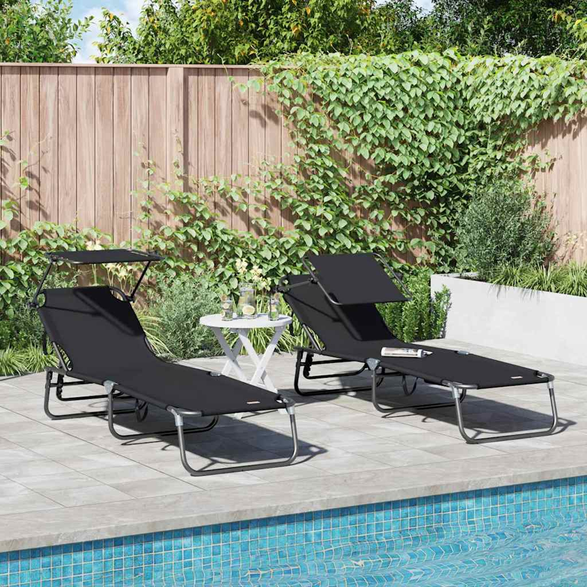 Zwei schwarze Liegestühle mit Sonnenschutz am Pool. Ein kleiner weißer Tisch hält Getränke. Ein Holzzaun und Grünzeug sind im Hintergrund.