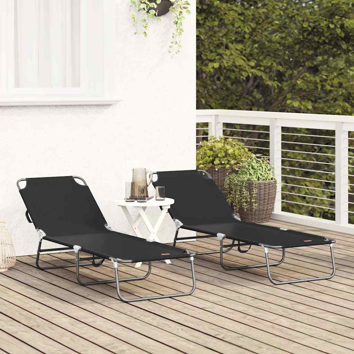 Zwei schwarze Liegestühle auf einer Holzterrasse, kleiner Tisch mit Getränken, grüne Pflanzen im Hintergrund und eine weiße Wand.