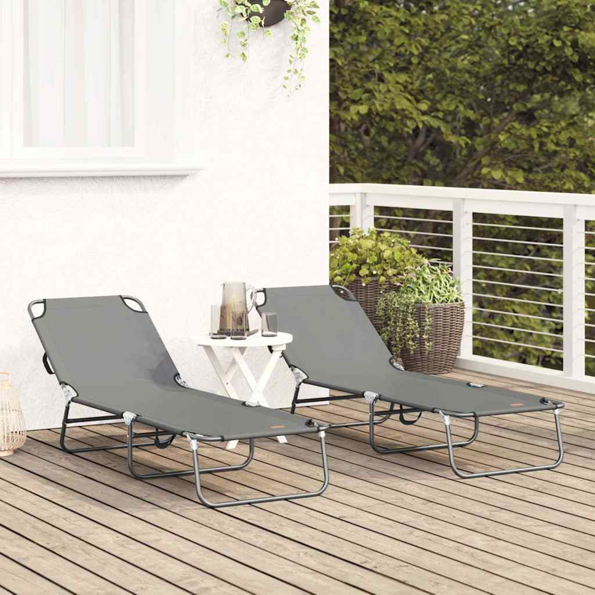 Zwei graue Liegestühle auf einem Holzdeck. Ein kleiner weißer Tisch trägt Gläser und einen Krug. Pflanzen und weißes Geländer im Hintergrund.