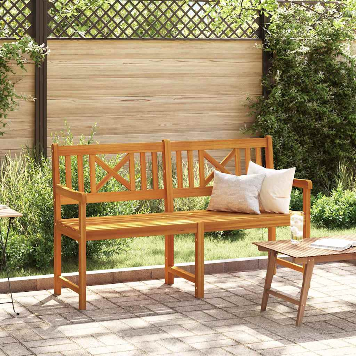 Un banco de madera con almohadas está en un patio. Una mesa pequeña con vaso y libro está a la derecha. Vegetación y valla en el fondo.