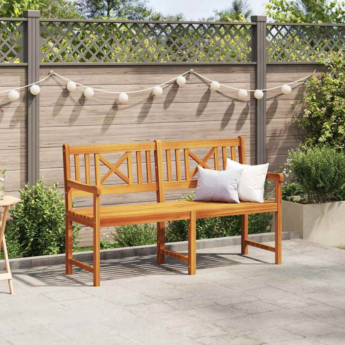 Banqueta de madera con almohadas en un jardín. Una valla con luces en el fondo. La vegetación exuberante rodea el banco.