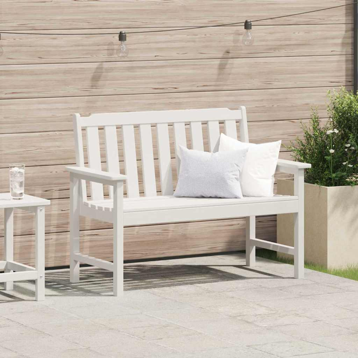 Panca bianca con cuscini su un patio. Un tavolino con un bicchiere d'acqua e luci decorative sono vicini. Parete in legno sullo sfondo.