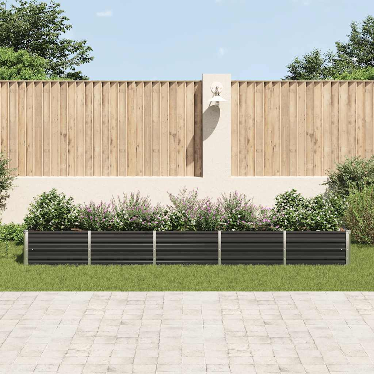 Eine Gartenszene mit einer schwarzen Pflanzkasten mit Blumen, vor einem Holzzaun und einer weißen Wand. Grünes Gras und Pflastersteine.