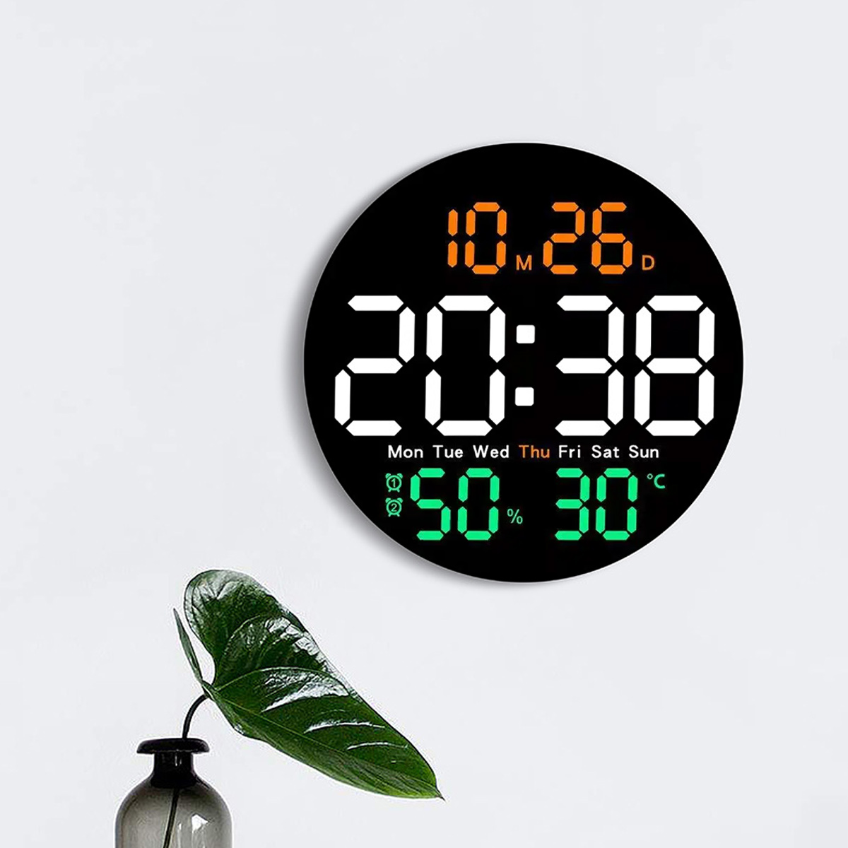 Eine Digitaluhr zeigt Datum und Uhrzeit, mit Luftfeuchtigkeit und Temperatur. Ein grünes Blatt in einer Vase.