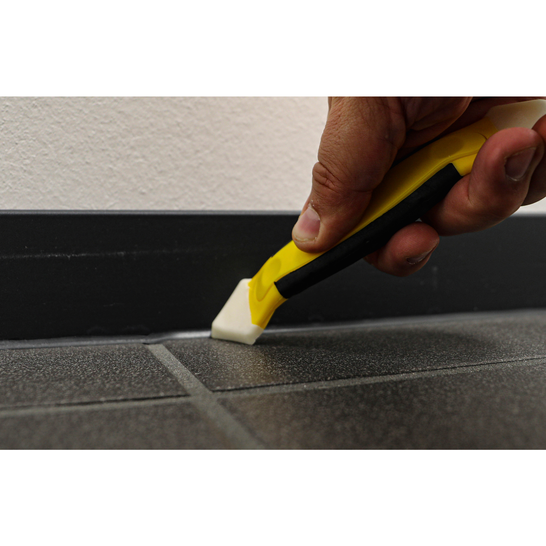 Una mano usa uno strumento giallo e nero per lisciare il mastice bianco lungo un pavimento scuro e un battiscopa nero, vicino a un muro bianco.