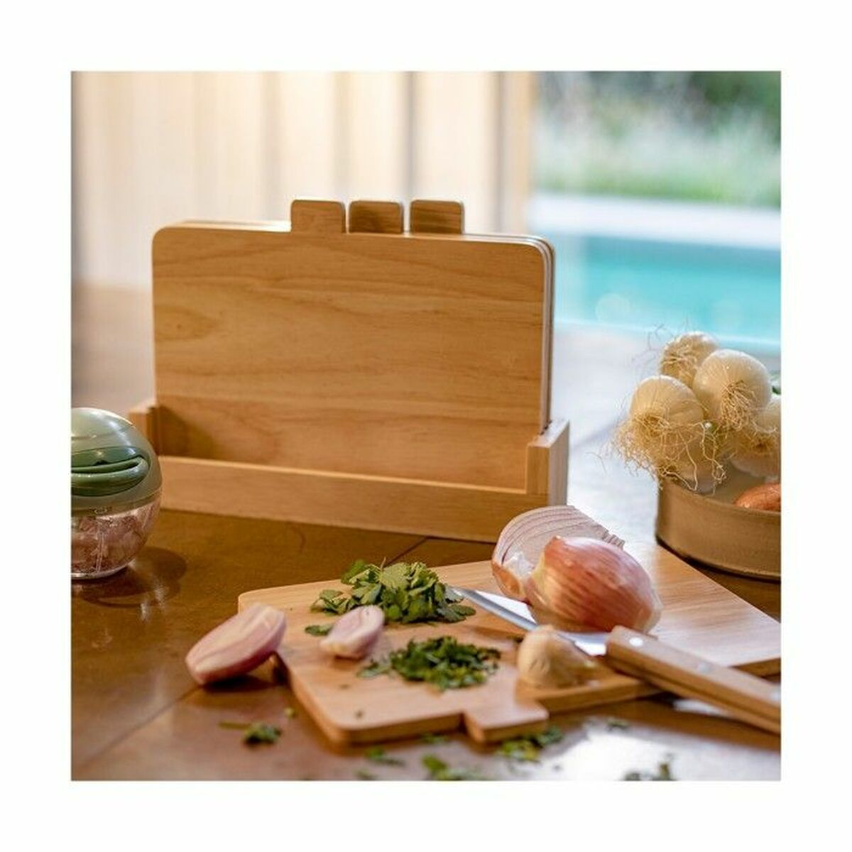 Holzschneidebretter mit gehackten Kräutern und Zwiebeln. Knoblauch und ein grüner Zerkleinerer sind sichtbar. Hintergrund zeigt Pool.